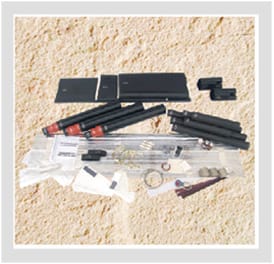 A GPS/2436 kit laid out on sand: four flat black joint-case plates, black and red-ended heat-shrink tubes, coiled mesh tape and spring clamps, mastic strips and sealed sachets.